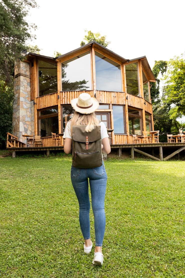 Mulher caminhando em direção a casas de madeira pré-fabricadas com mochila e chapéu, em ambiente arborizado.